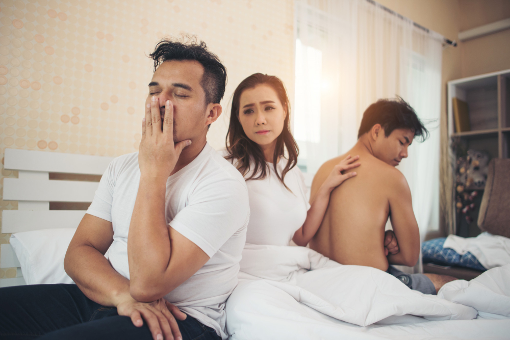 A man covering face in shame while sitting on a bed, a concerned woman touches his back suspecting he catch cheating spouse behind them, looking away and covering his private